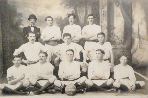 Ein Schwarz-Weiß-Foto einer Fussballmannschaft, mit einigen Spielern sitzend und einigen stehend, einem Ball in der Mitte und Bäumen mit einem klaren Himmel im Hintergrund, mit Text unten.