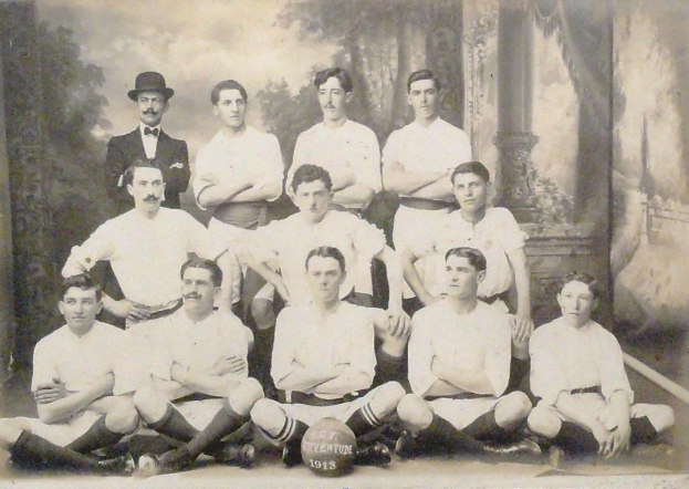 Ein Schwarz-Weiß-Foto einer Fussballmannschaft, mit einigen Spielern sitzend und einigen stehend, einem Ball in der Mitte und Bäumen mit einem klaren Himmel im Hintergrund, mit Text unten.