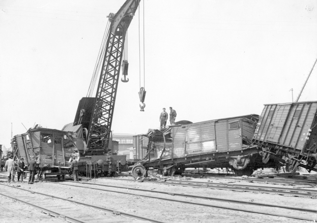 Schwarzes und weißes Bild von einer Zugkatastrophe mit einem Kran, der ein Waggon auf die Schienen hebt, mehrere Menschen in der Nähe, Gebäude und ein klarer Himmel im Hintergrund.