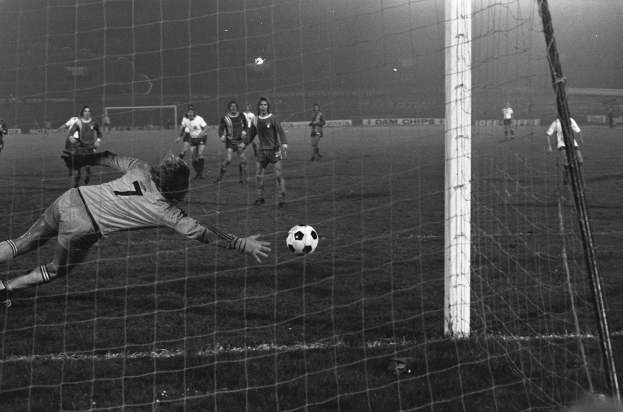 Ein Schwarz-Weiß-Foto eines Fußballspielers bei einer Parade vor einem Tor, umgeben von einer Gruppe von Menschen, die in der Nähe stehen und ein Torpfosten auf der rechten Seite sowie Werbetafeln und Lichter im Hintergrund zu sehen sind.