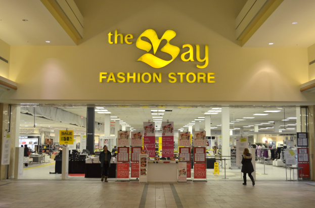 Außenansicht eines Bay Fashion Stores in einem Einkaufszentrum mit Menschen, Texttafeln an Wänden, Säulen, Deckenleuchten und Hintergrundauslagen.