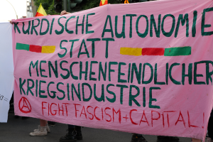 Eine Gruppe von Menschen steht auf einer Straße und hält ein pinkes Banner hoch, auf dem "Kurdische Autonomie Statt Menschenfeindlicher Kriegsindustrie Fight Fascism Capital" steht. Im Hintergrund sind Verkehrsschilder, Flaggen, Bäume, Gebäude und ein klarer blauer Himmel zu sehen.