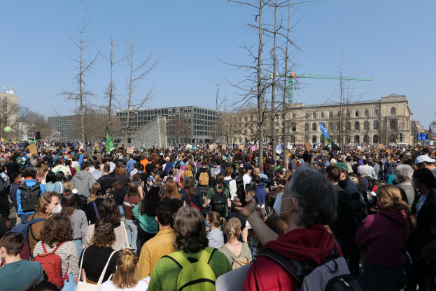 Eine große Menge Menschen mit Schildern und Taschen vor einem Gebäude mit Fenstern, Bäumen und einem klaren Himmel während einer Klimaprotest