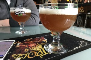 Zwei Gläser goldenen Biers auf einem Tisch neben einem Handy, mit Menschen und gerahmten Wanddekorationen im Hintergrund.