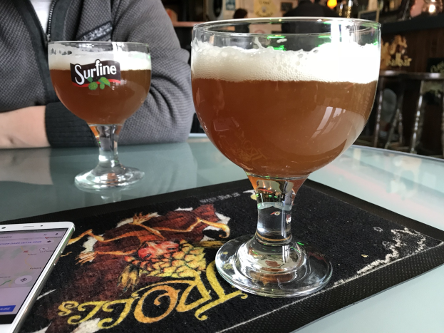 Zwei Gläser goldenen Biers auf einem Tisch neben einem Handy, mit Menschen und gerahmten Wanddekorationen im Hintergrund.