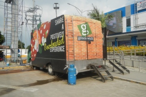 Ein großer Lastwagen steht vor einem Gebäude mit einer Tafel, auf der "Teilen Sie eine positiv überladene Erfahrung" steht, mit Stufen, einem Zylinder auf dem Boden, Geländern, Strommasten mit Drähten, Laternen, Bäumen und einem bewölkten Himmel im Hintergrund.