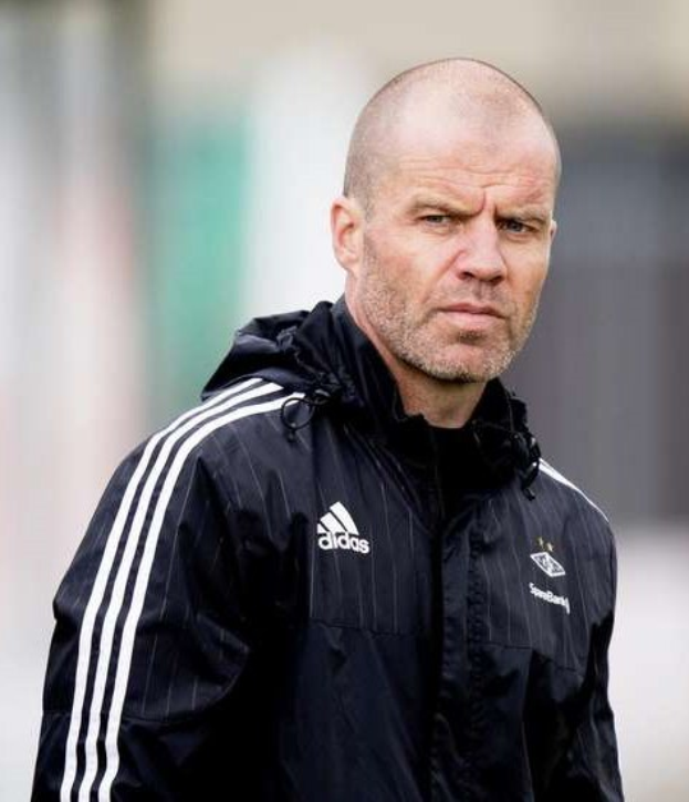 Ein kahlköpfiger Trainer in einer blauen Jacke mit weißen Streifen und einem weißen Kragen steht auf einem Fußballfeld mit gefalteten Händen, ernster Gesichtsausdruck, unscharfer Hintergrund.