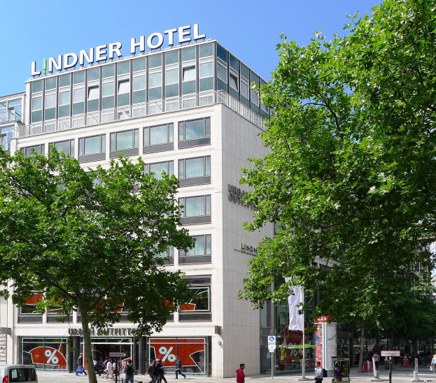 Das Lindner Hotel in Berlin, ein großes Glasfrontgebäude mit Beschriftung oben drauf, umgeben von Bäumen und einer belebten Straße mit Fußgängern und Fahrzeugen.