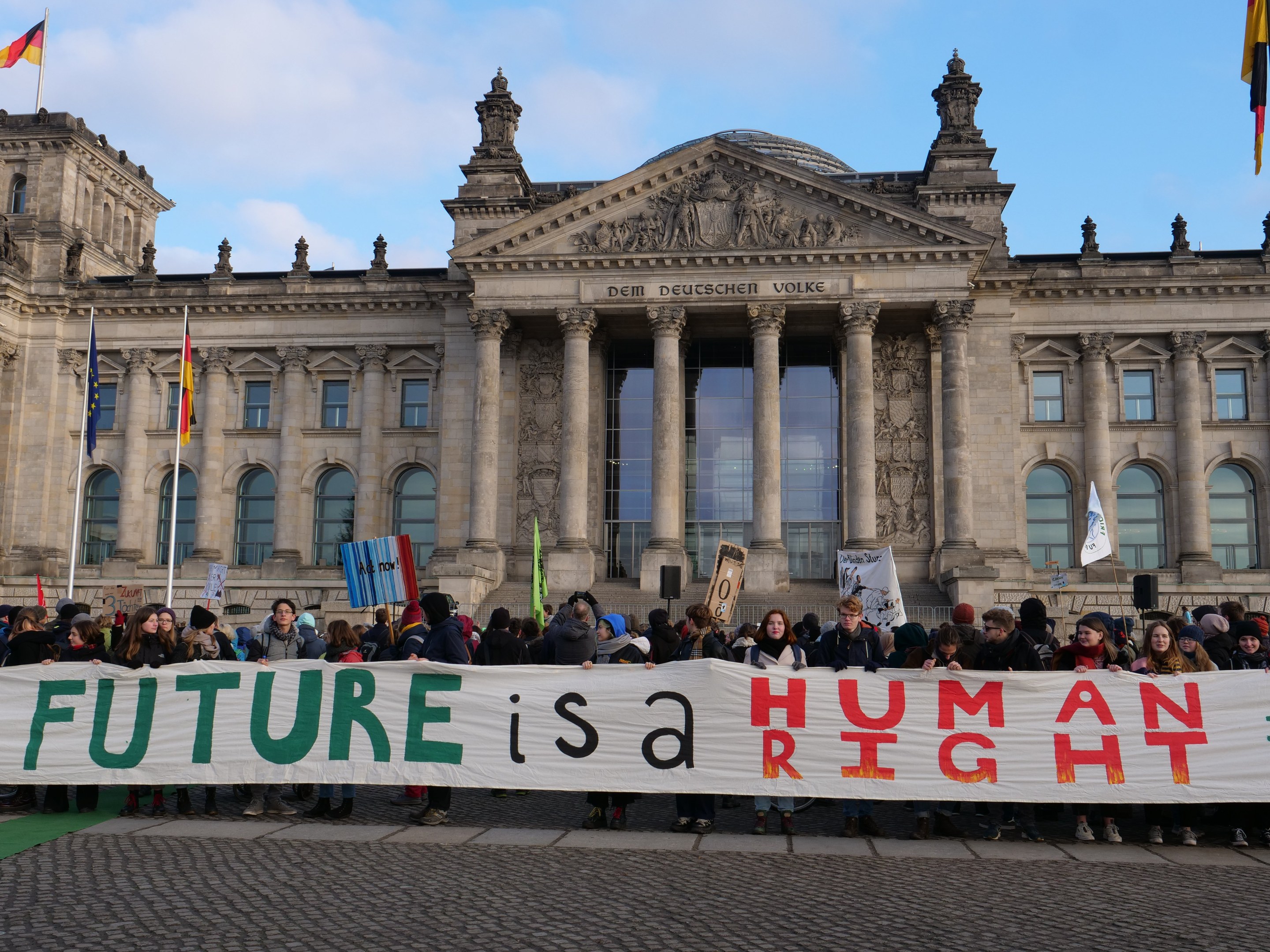 Eine Gruppe von Menschen mit einem Transparent 'Zukunft ist ein Menschenrecht' vor dem Reichstagsgebäude in Berlin, Deutschland, mit sichtbaren Fenstern, Säulen und Bögen des Gebäudes, umgeben von Fahnenmasten und einem bewölkten Himmel.