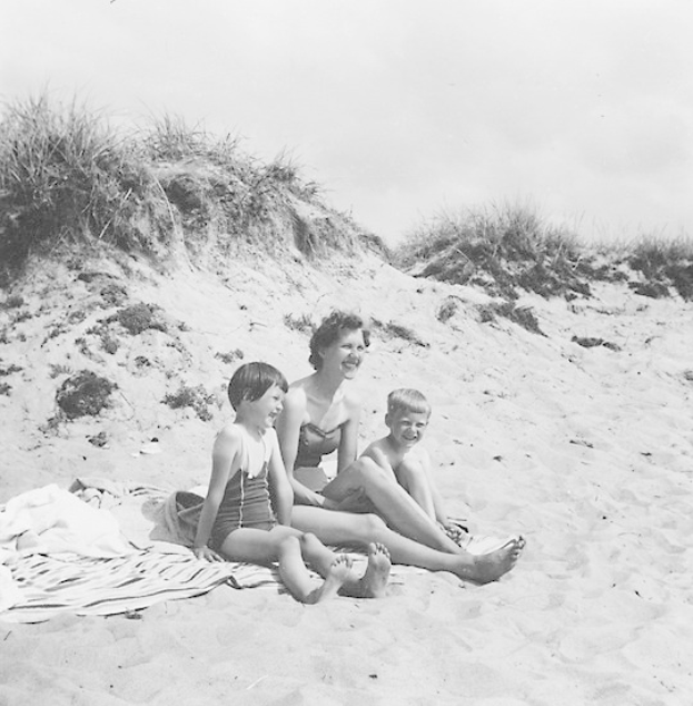 Schwarz-weißes Foto einer Frau in einem Bikini und zwei Kindern in Badebekleidung, die auf einer Decke am Strand sitzen, mit Sand, Gras und Himmel im Hintergrund.