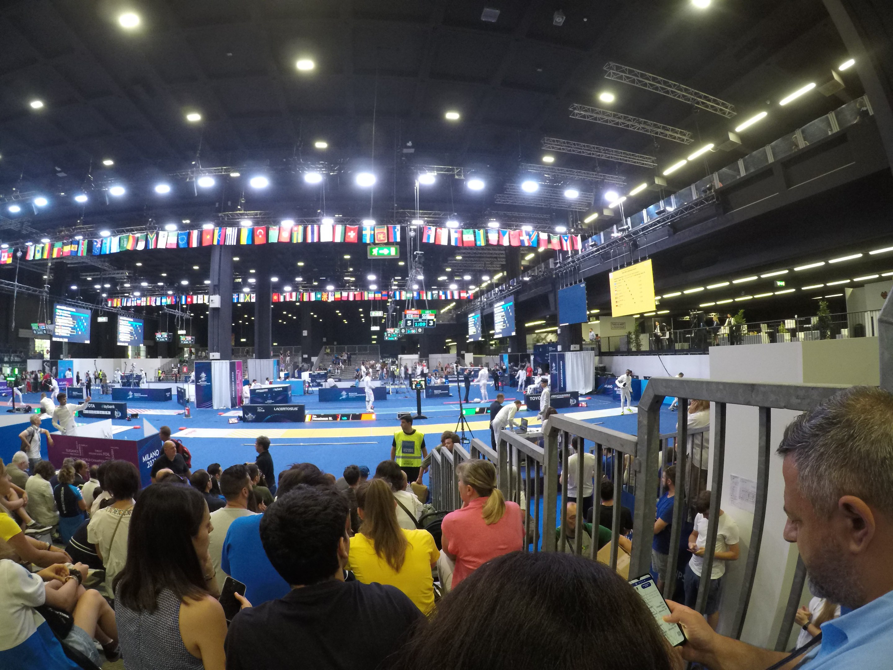 Große Menschenmenge auf Sitzplätzen und Stehplätzen in einer Indoor-Sportarena mit Bildschirmen, Flaggen und Deckenbeleuchtung.