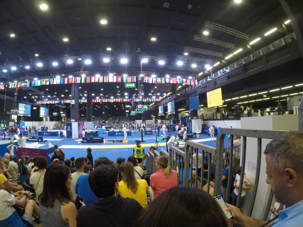 Große Menschenmenge auf Sitzplätzen und Stehplätzen in einer Indoor-Sportarena mit Bildschirmen, Flaggen und Deckenbeleuchtung.
