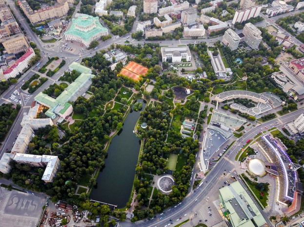 Luftaufnahme einer Stadt mit dichtem Baumbestand, Straßen, Fahrzeugen und Grünflächen, mit einem zentralen Park, der von Bäumen und Vegetation umgeben ist.
