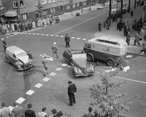 Schwarz-weißes Bild eines Autounfalls an einer Straßenkreuzung mit mehreren Fahrzeugen, darunter ein Van, und einer Gruppe von Schaulustigen; Gebäude, Bäume, Strommasten und Texttafeln sind im Hintergrund zu sehen.