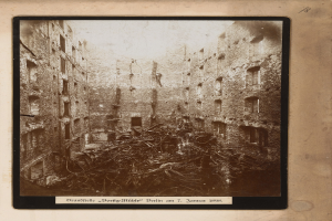 Altes Foto eines zerstörten Gebäudes in einer städtischen Umgebung, gerahmt mit Schutt und zerbrochenen Fenstern, und Text unten.