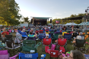 Eine große Menschenmenge sitzt auf Klappstühlen vor einer Bühne beim Nelsonville Music Festival, umgeben von Bäumen und Himmel, ein paar Menschen stehen rechts auf der Bühne, links ein Schuppen.