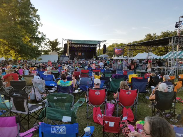 Eine große Menschenmenge sitzt auf Klappstühlen vor einer Bühne beim Nelsonville Music Festival, umgeben von Bäumen und Himmel, ein paar Menschen stehen rechts auf der Bühne, links ein Schuppen.