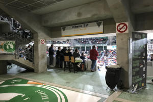 Gruppe von Menschen vor sitzendem Publikum in einem Stadion mit Schildern, die "Boston Celtics vs New York Jets" lesen, einem Müllcontainer, einer Tafel mit Text, einer Treppe mit Geländern und einer Decke mit Deckenleuchten.