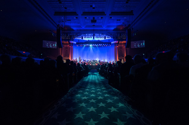 Ein großer Saal mit in Reihen sitzenden Menschen, die einer Bühne mit Musikern gegenüberliegen, unter einer Decke mit Lichtern und Fernsehgeräten, was auf eine Konzertsäleinrichtung hinweist.