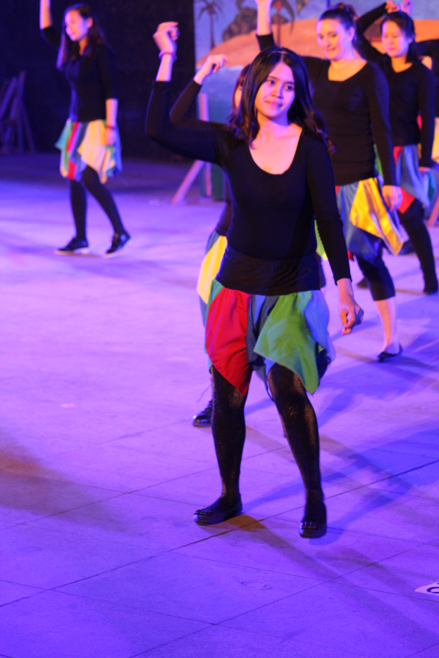 Eine Gruppe junger Frauen, die auf einer Bühne tanzen, mit einem Gemälde an der Wand im Hintergrund, alle tragen Schuhe.