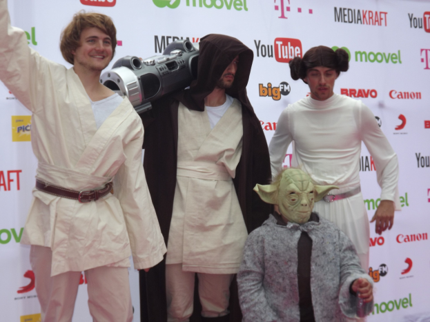 Eine Gruppe von Menschen in Star-Wars-Kostümen, die auf einem roten Teppich posieren, wobei eine Person ein Objekt und ein Banner mit Text und Logos im Hintergrund hält.