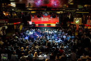 Eine große Menschenmenge sitzt auf Stühlen vor einer Bühne mit Bildschirmen und Bannern, die von Scheinwerfern beleuchtet wird und von Geländern umgeben ist und gespannt auf den Beginn eines Online-Marketing-Rockstars-Events wartet, mit einem Wasserzeichen in der linken unteren Ecke.