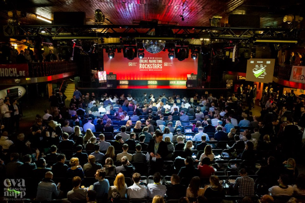 Eine große Menschenmenge sitzt auf Stühlen vor einer Bühne mit Bildschirmen und Bannern, die von Scheinwerfern beleuchtet wird und von Geländern umgeben ist und gespannt auf den Beginn eines Online-Marketing-Rockstars-Events wartet, mit einem Wasserzeichen in der linken unteren Ecke.