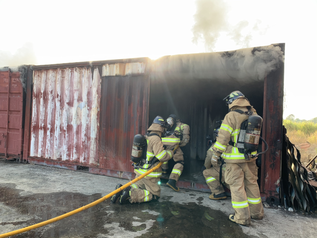 Feuerwehrleute in Schutzausrüstung löschen ein Feuer in einem Container, mit Rauchentwicklung und Grünzeug im Hintergrund.