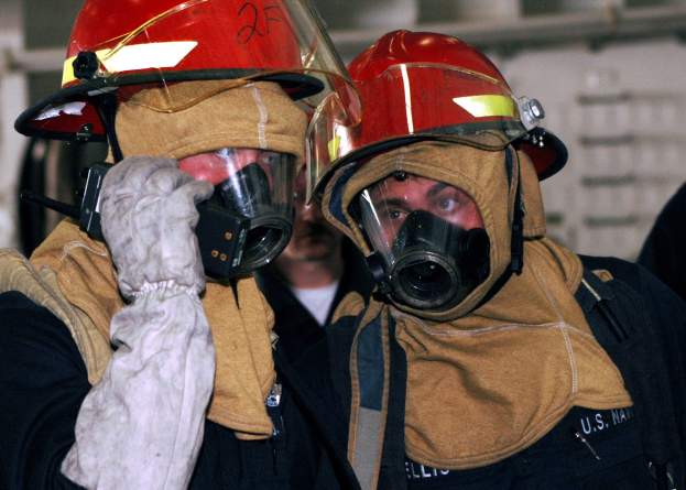 Zwei Feuerwehrleute in Gasmasken und Helmen, einer hält ein Funkgerät in der Hand, stehen neben einer Wand mit freiliegenden Rohren.