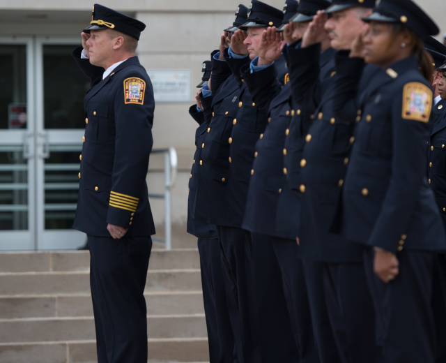 Eine Gruppe von Polizisten in Uniform und Mützen steht in Formation und salutiert vor einem Gebäude mit Glastüren und Treppen.