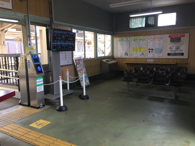 Ein belebter Bahnhofseingang mit Sitzgelegenheiten, einer Informations counter, Ticketautomaten, Kettenbarrieren, Informationsanzeigen, Wandplakate, Deckenbeleuchtung und Geländern.