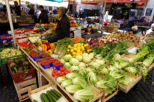 Ein belebter Outdoor-Markt mit einer Vielfalt an Obst und Gemüse, Menschen unter Zelten sitzend und stehend und Gebäude mit Text im Hintergrund.