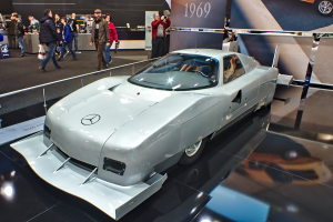 Ein silberner Mercedes-Benz 300 SL Gullwing wird auf der Frankfurter Motor Show präsentiert, umgeben von Menschen in verschiedenen farbigen Kleidern, mit Schildern und anderen Gegenständen im Hintergrund.