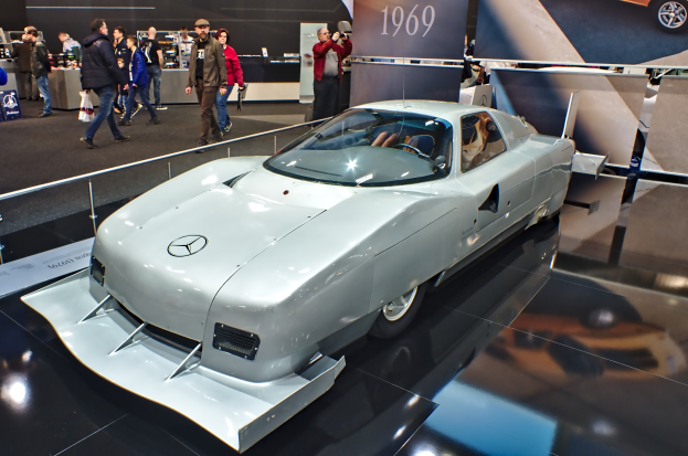 Ein silberner Mercedes-Benz 300 SL Gullwing wird auf der Frankfurter Motor Show präsentiert, umgeben von Menschen in verschiedenen farbigen Kleidern, mit Schildern und anderen Gegenständen im Hintergrund.