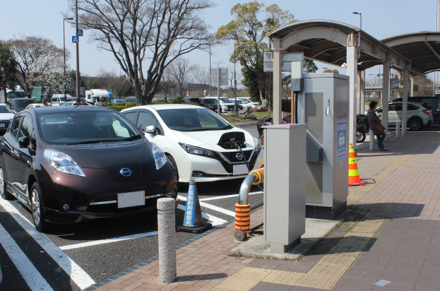 Elektroauto-Ladestation in Japan mit Autos, Verkehrskegeln, einer Person auf dem Gehweg, einem Schuppen, Polen, Lampen, Schildern, Bäumen, Pflanzen und einem Himmel im Hintergrund.