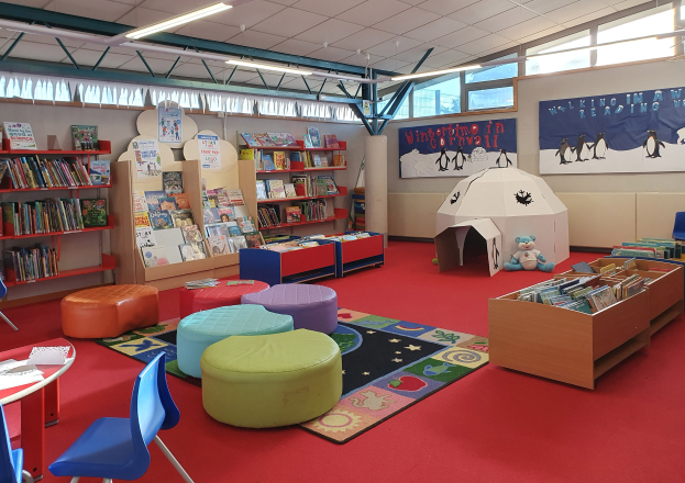 Eine hell erleuchtete Kinderbibliothek mit Bücherregalen, Tischen, Stühlen, Hockern, Teppichen, Wandbrettern, Deckenleuchten und großen Fenstern.