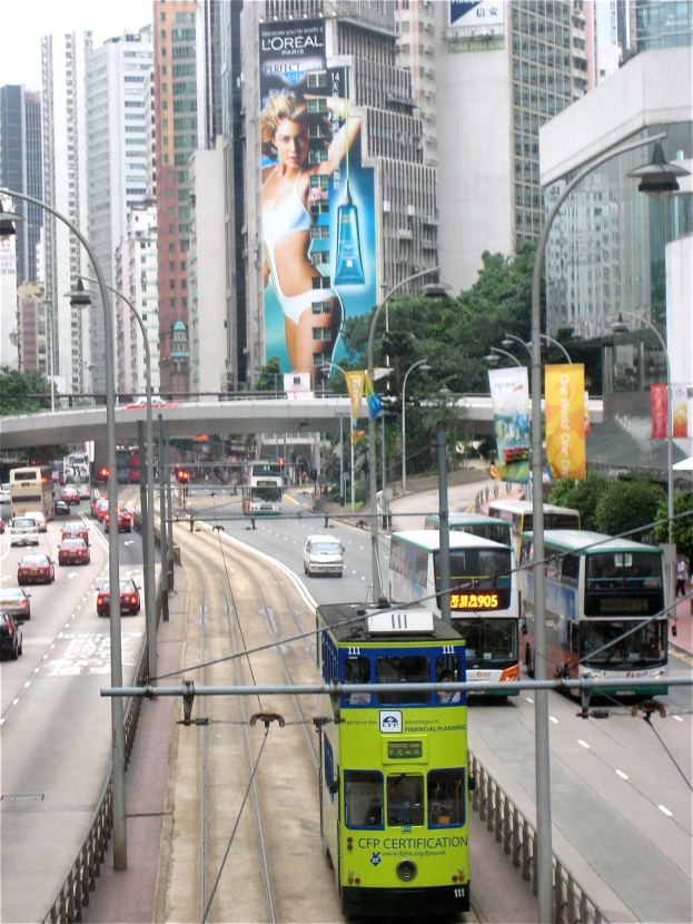 Eine belebte Stadtstraße mit verschiedenen Fahrzeugen, darunter Doppeldeckerbusse, Autos und andere Fahrzeuge, unter einem klaren blauen Himmel, mit hohen Gebäuden, einer Brücke und Text unten.