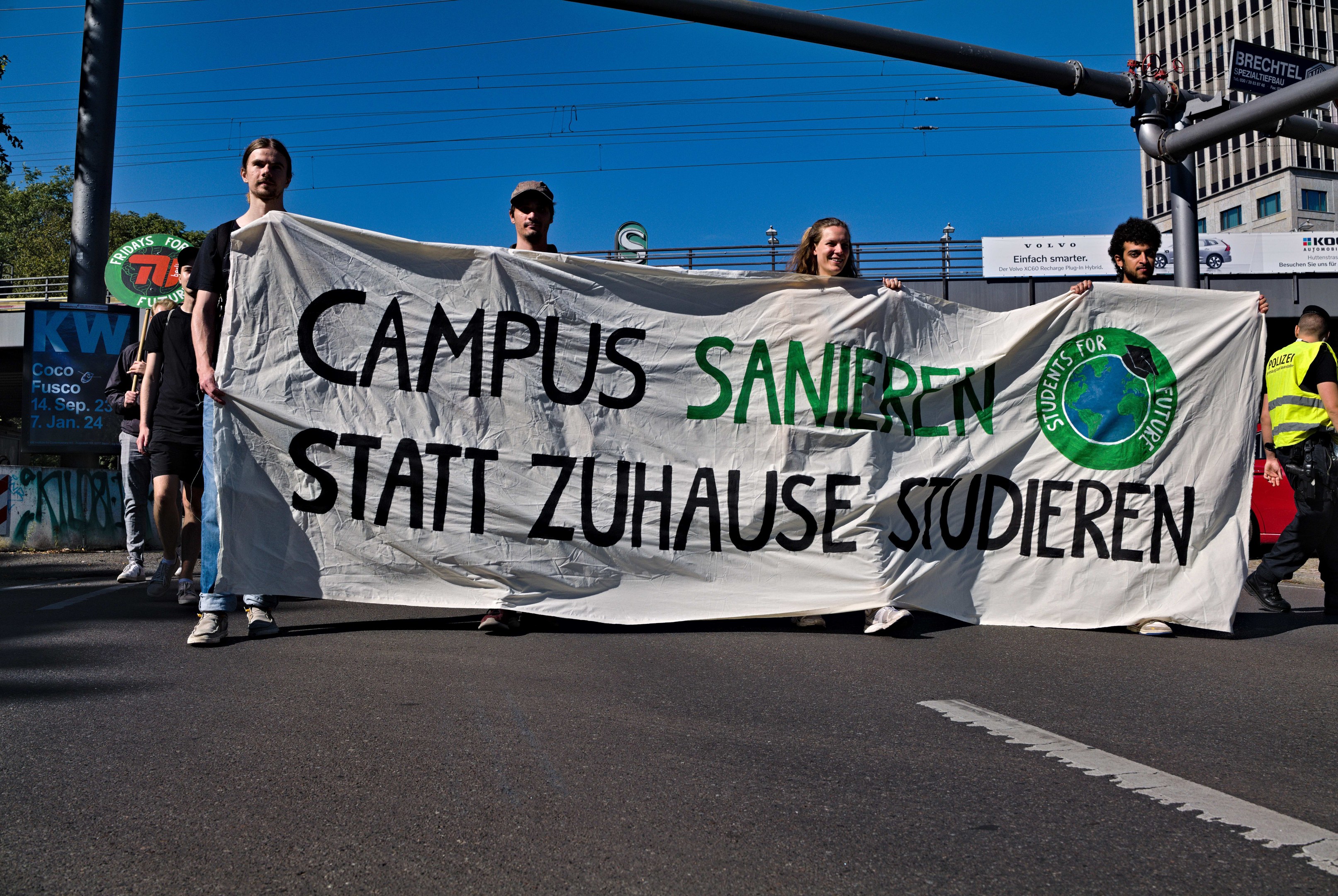 Eine Gruppe von Menschen geht mit einem Transparent, auf dem "Campus Sanien Statt Zuhause Studieren" steht, eine Straße entlang, mit Gebäuden, Bäumen und einem klaren blauen Himmel im Hintergrund.