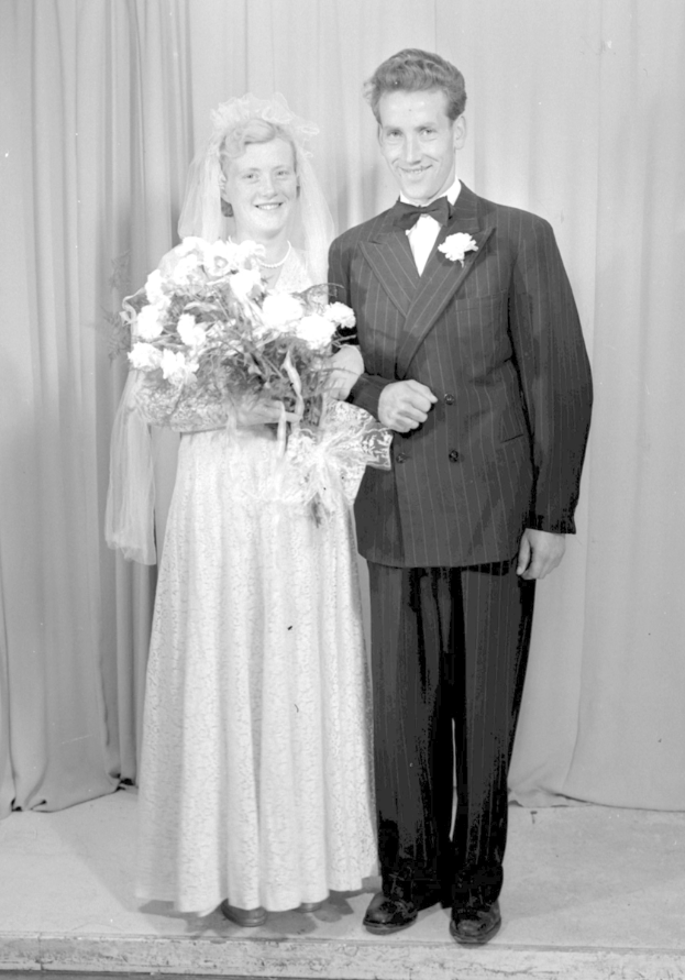 Ein Mann und eine Frau stehen nebeneinander auf einer Bühne, lächeln und die Frau hält einen Blumenstrauß, während ein Vorhang hinter ihnen zu sehen ist.