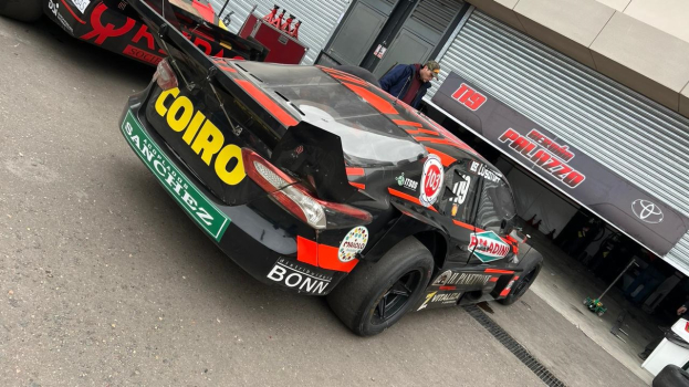 Eine Gruppe von Autos steht vor einem Gebäude mit Ladenschutz und einem Banner mit der Aufschrift "Le Mans 2019", umgeben von Menschen, was den Start eines Rennens vermuten lässt.