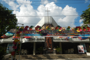 Ein Gebäude mit einer Tafel, die "Frohes Fest St. Josef der Arbeiter" anzeigt, davor stehend, mit einer Treppe zum Eingang, einem Bogen mit hängenden Dekorationen, Außenbestuhlung, Topfpflanzen, Bäumen und einem bewölkten Himmel.