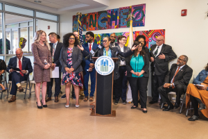 Gruppe von Menschen vor einem Podium mit Mikrofon und einer Tafel mit der Aufschrift "Sancho-Stadtrat tritt dem Stadtrat bei", mit sitzenden und stehenden Teilnehmern, Flaggen, einem Feuerlöscher und Deckenleuchten im Hintergrund.