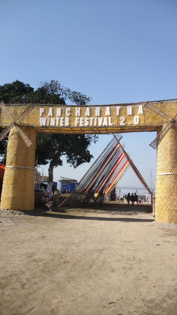 Ein festliches Bild mit einem Riesenrad in der Mitte, einem Tor mit Text, einem Zaun, einer Gruppe von Menschen auf dem Boden, einigen Pfählen, einem Turm, Bäumen und einem bewölkten Himmel.