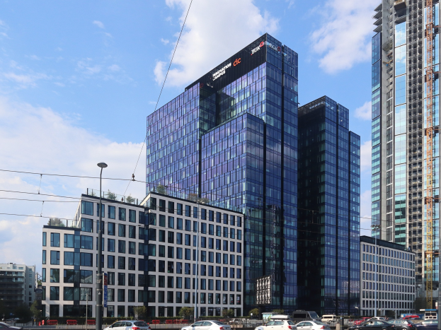 Großes Gebäude mit vielen Fenstern, identifiziert als Deutsche Bank Hauptsitz in Frankfurt, Deutschland, umgeben von städtischer Infrastruktur einschließlich Straßenlaternen, Fahrzeugen, Grünflächen und einem bewölkten Himmel.
