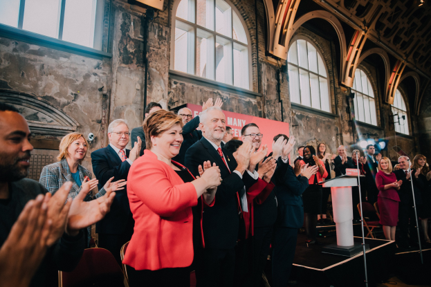 Eine Gruppe von Menschen, die vor einer Menge klatschen, mit einem Podium, einem Mikrofon, Stühlen, einer Wand, Fenstern und einem Banner mit der Aufschrift "Covid-19 New York City Mayor Bill de Blasio" im Hintergrund.