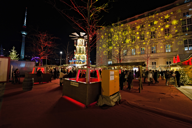 Ein lebendiger Weihnachtsmarkt in Berlin, Deutschland, mit Menschen um geschmückte Stände, Bäume, Gebäude, Laternenmasten und einen Turm unter einem dunklen Himmel.