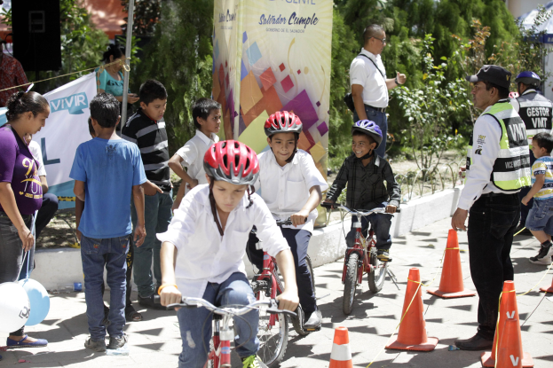Gruppe von Kindern, die auf der Straße mit Verkehrsleitkegeln Fahrrad fahren, einige tragen Helme, andere stehen in der Nähe, mit einem Banner, Bäumen und Gebäuden im Hintergrund.