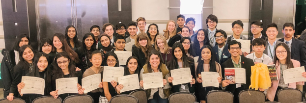 Gruppe von Menschen mit Urkunden, die für ein Foto in einem Raum mit Stühlen und einer Wand im Hintergrund posieren, wahrscheinlich bei einer Abschlussfeier.