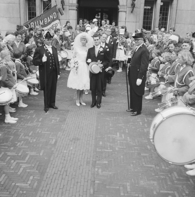 Schwarzes und weißes Foto einer Braut in einem weißen Kleid und Schleier und eines Bräutigams mit einem Strauß, der eine Straße entlang geht, vorbei an einer Menge mit Musikinstrumenten, mit einem Banner und einem Gebäude im Hintergrund.