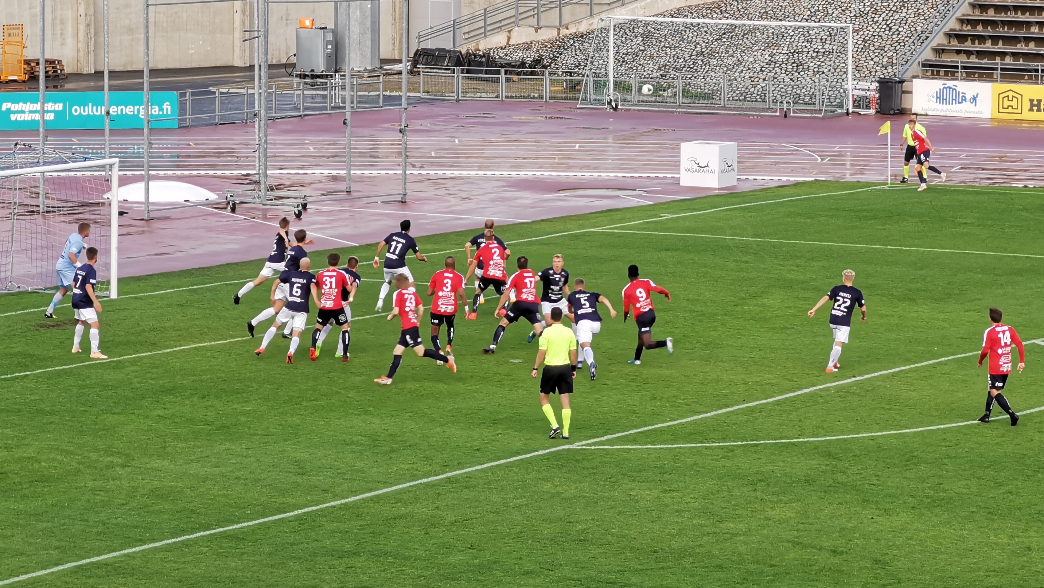 Gruppe von Menschen, die Fußball auf einem Feld mit Zuschauern in Stadionrängen, umgeben von Pfählen, Schildern mit Text, Geländern, Stufen und anderen Gegenständen spielen.